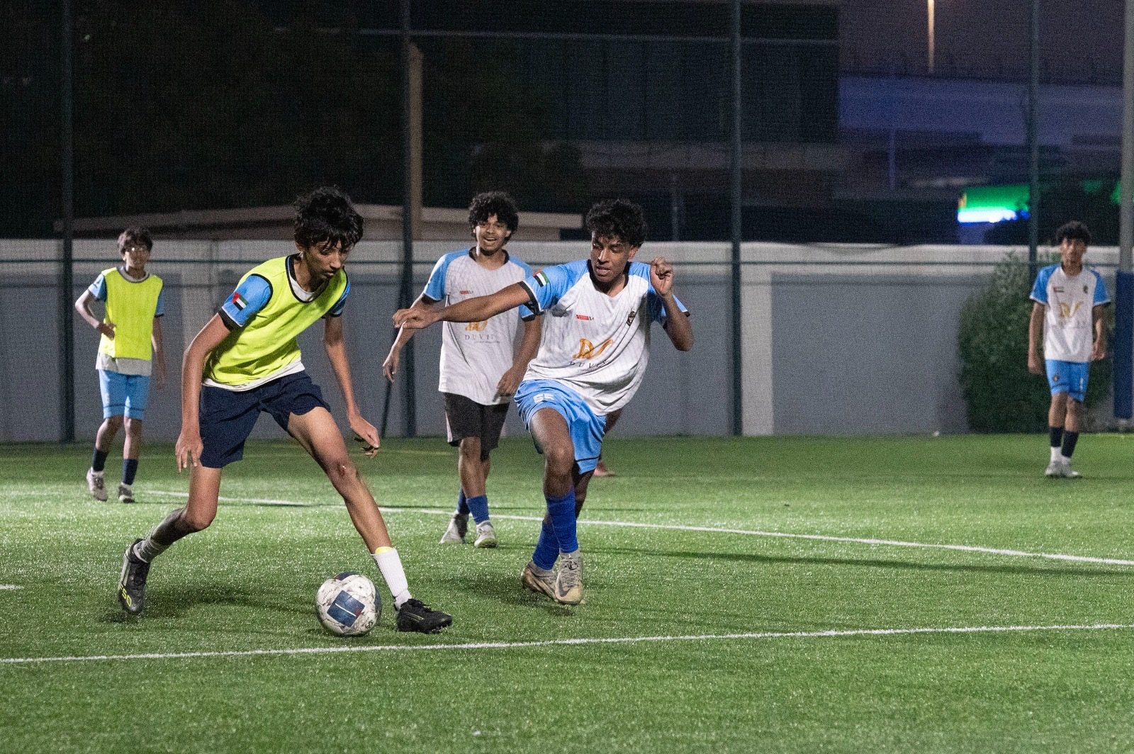 Dev dribbling past a defender during a floodlit night match